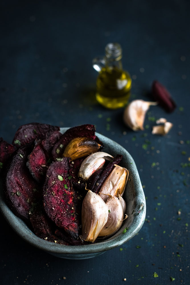 tigela com beterrabas e dentes de alho assados em primeiro plano, com ingredientes ao fundo.